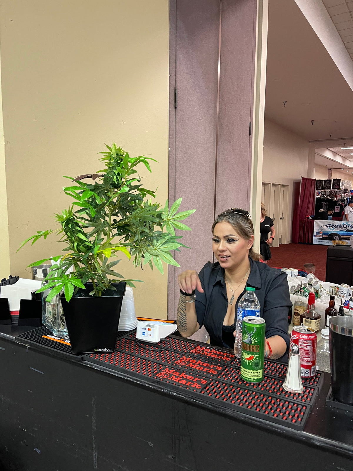 Woman at a bar talking with a DecoBudz faux cannabis plant beside her, styled as bold statement decor