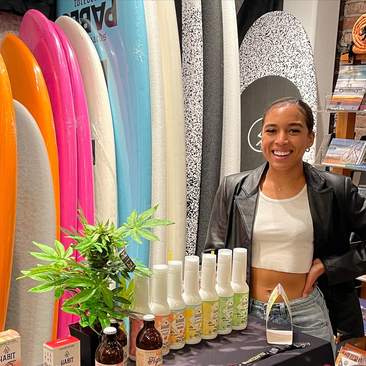 DecoBudz faux cannabis plants displayed on a table showcasing CBD products at a trade show, with surfboards in the background